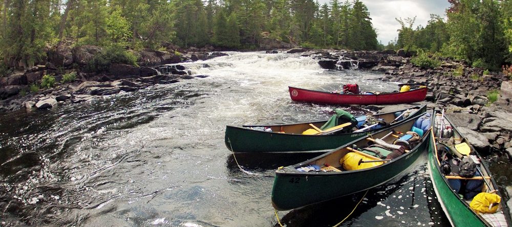 Canoes tied to shore with whitewater in background HEADER 1000x445