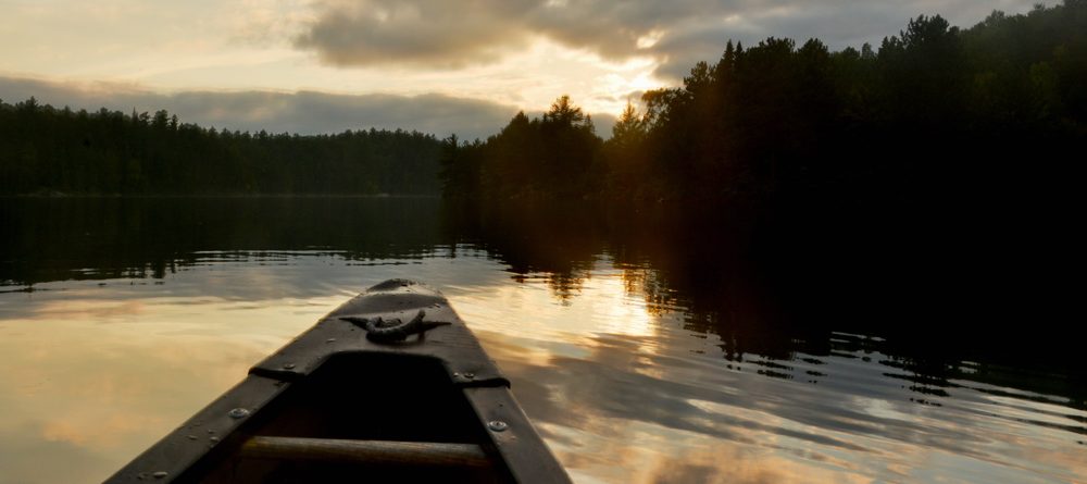 Front of a canoe on a lake in with the sun setting 1000x445