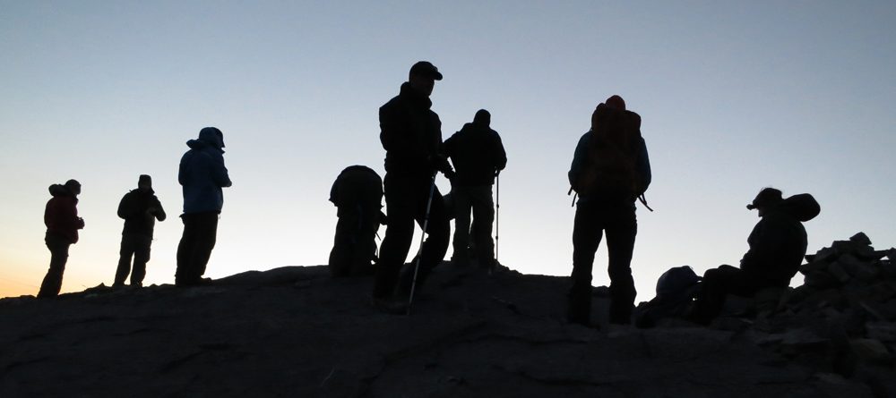 Silhouette of veterans in mountaineering gear standing on rock with blue sky behind Header 1 1000x445