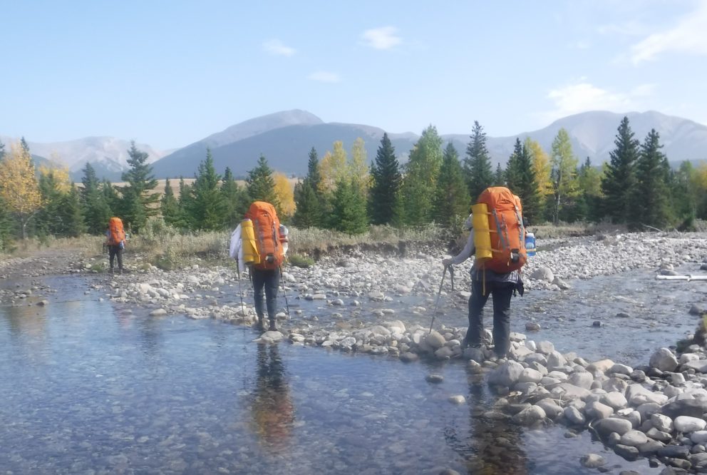 Three people crossing rocky stream with backpacks, mountains and trees in the distance
