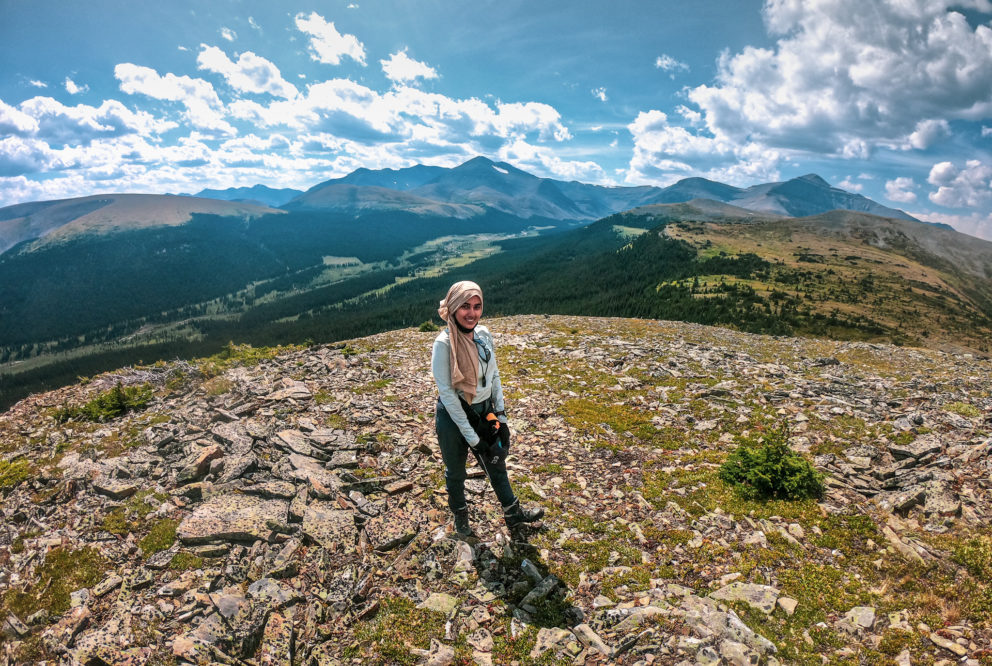 Image of person standing at the top of a mountain overlooking a valley surrounded by moutain peaks.