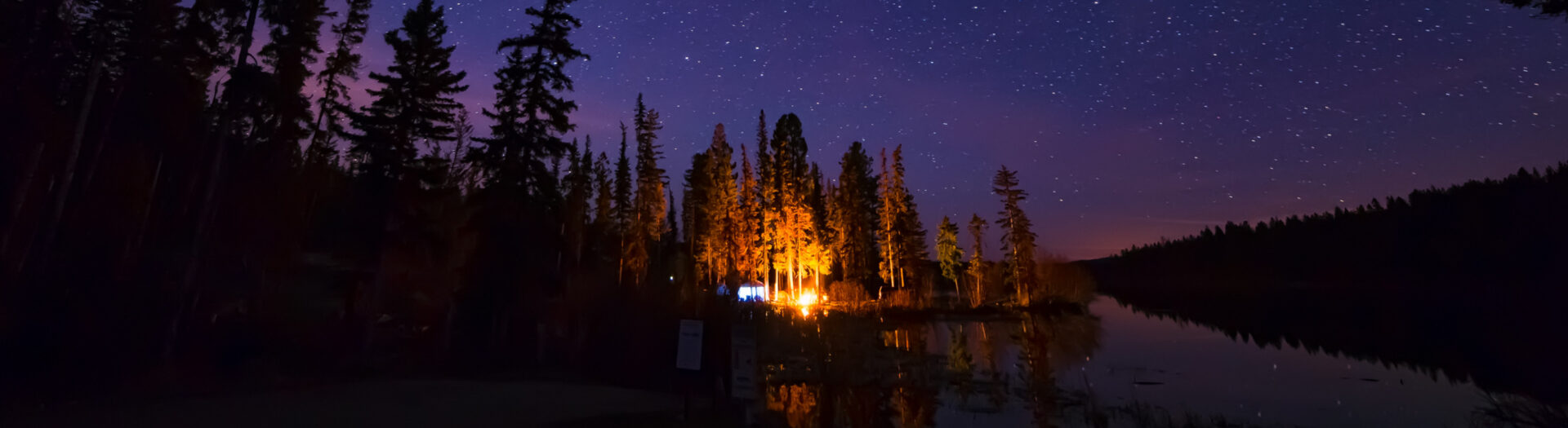 Image of a campfire at night next to a still lake with a starry sky above